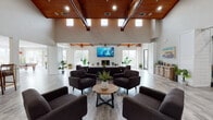 This single-story space makes a striking first impression through its dramatic vaulted ceilings clad in warm wood paneling, supported by exposed timber beams that lend an architectural richness to the interior. The main gathering area is expansive and light-filled, drawing natural light through a row of clerestory windows positioned high along the roofline, as well as through large windows that frame views of the surrounding greenery. White shiplap walls contrast beautifully with the warm wood ceiling and gray wood-look flooring that flows throughout the space.

The primary lounge area is anchored by a fireplace and wall-mounted flat-screen television, with generous seating arrangements of charcoal upholstered armchairs grouped around a round wood coffee table. The open layout allows for flexible furniture configurations across the broad floor plan. A secondary lounge near the entry features a classic barn door and plantation shutters, adding to the coastal-inspired character.

The fully equipped kitchen features white shaker cabinetry, gray countertops, white subway tile backsplash, and stainless steel appliances including a refrigerator, range, and microwave. A peninsula counter connects the kitchen to the main living area. The 3D floor plan also reveals a dedicated game room with a pool table, completing this versatile and welcoming space.