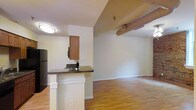 This single-floor unit offers a practical and comfortable layout centered around an open-concept living and kitchen area. The kitchen is equipped with dark-finished cabinetry, black appliances including a refrigerator and microwave, and a peninsula counter finished with a dark granite-style surface — ideal for casual dining or food preparation. The peninsula features decorative column accents that add a touch of architectural character to the space.

Warm hardwood-style flooring runs throughout the main living areas, creating a cohesive and inviting atmosphere. The open layout allows the kitchen to flow seamlessly into the adjacent living area, which is generously sized and bathed in a neutral color palette of soft whites and warm beige tones. Flush-mount ceiling lighting provides consistent illumination throughout.

The entry to the unit is framed by a classic white paneled door, providing a welcoming first impression. Exposed ductwork is visible along the ceiling of the living area, lending the space a subtle industrial character. Clean white baseboards and neutral walls offer a versatile backdrop suitable for a variety of interior styles. Overall, the space combines functional design with a warm, approachable ambiance.