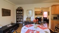 Unit A302 is a single-floor residence that presents a comfortable and well-utilized layout, with the dining area flowing naturally into the living space beyond. The dining area serves as a central gathering point, furnished with a round table dressed in a decorative white and red embroidered tablecloth, surrounded by metal-frame chairs. A floral centerpiece adds a warm, inviting touch to the setting.

A tall, dark wood bookcase dominates one wall of the dining area, filled with an extensive collection of books, DVDs, and media, lending the space a lived-in, intellectual character. Beside it, a lower wooden console holds decorative pieces and collectibles, contributing to the room's layered, personal aesthetic.

Through an open passageway, the living area is visible, featuring a landscape painting as a prominent wall accent, a television setup, and additional seating. Warm lighting and pink-toned curtains in the background give the living space a cozy ambiance. The hardwood flooring runs throughout, unifying the areas cohesively. Overall, the space offers a functional and characterful single-level layout.