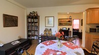Unit A302 is a single-floor residence that presents a comfortable and well-utilized layout, with the dining area flowing naturally into the living space beyond. The dining area serves as a central gathering point, furnished with a round table dressed in a decorative white and red embroidered tablecloth, surrounded by metal-frame chairs. A floral centerpiece adds a warm, inviting touch to the setting.

A tall, dark wood bookcase dominates one wall of the dining area, filled with an extensive collection of books, DVDs, and media, lending the space a lived-in, intellectual character. Beside it, a lower wooden console holds decorative pieces and collectibles, contributing to the room's layered, personal aesthetic.

Through an open passageway, the living area is visible, featuring a landscape painting as a prominent wall accent, a television setup, and additional seating. Warm lighting and pink-toned curtains in the background give the living space a cozy ambiance. The hardwood flooring runs throughout, unifying the areas cohesively. Overall, the space offers a functional and characterful single-level layout.