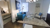 This single-floor space showcases a thoughtfully designed living area that blends contemporary style with a curated selection of furnishings and décor. The room is anchored by rich hardwood flooring, which provides a warm contrast to the cool, neutral-toned walls. A striking teal velvet sofa serves as the focal point, complemented by a plush cream area rug and a sleek acrylic and glass coffee table that adds a sense of openness to the space.

Overhead, a multi-globe spherical chandelier introduces a mid-century modern aesthetic, casting ambient light throughout the room. An oversized bold blue geometric canvas artwork on one wall adds a graphic, artistic element, while a subtler abstract painting on the adjacent wall offers visual balance. Natural light filters through a window fitted with horizontal blinds, contributing to the bright, airy atmosphere.

Accent pieces—including a lucite side table, a sculptural floor lamp, and a marble-topped side table—demonstrate careful attention to detail. A classic accent chair with dark wood legs rounds out the seating arrangement, creating an inviting, well-proportioned layout ideal for both relaxing and entertaining.