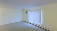 This single-floor space offers a clean, understated layout designed for functional living. The interior is finished with soft neutral walls and wall-to-wall carpeting in a light gray tone, creating a calm and cohesive atmosphere throughout. The main living area is notably open, providing a comfortable sense of space and ease of movement.

A standout practical feature of the space is its dual climate control system: a wall-mounted air conditioning unit ensures year-round cooling, while a continuous baseboard heating unit runs along the base of the exterior wall for efficient warmth during colder months. This combination makes the space adaptable to a range of seasonal conditions.

Natural light is a key characteristic of the interior, with a large window dressed in vertical blinds that allows for adjustable light control and a bright, airy feel. When the blinds are open, the window floods the room with natural light, enhancing the overall sense of openness in the space.

The overall design is straightforward and well-maintained, with clean white ceilings and smooth walls that contribute to a bright and tidy presentation. The minimalist aesthetic allows the space to be easily personalized, making it a practical and versatile option for a variety of uses.