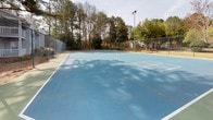 This outdoor recreational space centers around a well-defined tennis court finished in a two-tone blue and green hard surface, with crisp white court markings clearly delineating the playing area. The court is fully enclosed by sturdy green chain-link fencing, providing both safety and a sense of containment for active play. Overhead court lighting is installed on tall poles, allowing for extended use during evening hours.

The facility is set within a naturally wooded environment, with mature pine and deciduous trees lining the perimeter and casting dappled shadows across the court surface. The surrounding landscape features a mix of mulched ground cover and natural pine straw, lending a relaxed, park-like atmosphere to the setting. A seating area with benches is visible near the court entrance, offering a convenient spot for players to rest between matches.

Adjacent to the main court area is a secondary surface in a sage green finish, which appears to serve as an additional playing or practice zone. The broader surroundings include nearby multi-story residential buildings with balconies and a small playground visible in the background, suggesting this court is part of a larger community amenity area. The open-air setting, mature tree canopy, and well-maintained fencing combine to create an inviting and functional recreational environment.