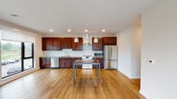 This well-appointed single-floor unit offers a thoughtfully designed open-concept layout that seamlessly connects the kitchen and living areas. The kitchen is a standout feature, showcasing rich dark wood cabinetry that extends to the ceiling for generous storage, a full suite of stainless steel appliances including a French door refrigerator, electric range, built-in microwave, and dishwasher, complemented by a sleek wall-mounted range hood and pendant lighting above a central island. Light hardwood flooring runs throughout the main living area, enhancing the bright, airy atmosphere created by large windows and recessed lighting. The spacious living room opens directly onto a private balcony through floor-to-ceiling sliding glass doors, offering open outdoor views. The bedrooms are carpeted for comfort and feature large windows with roller shades, allowing ample natural light while maintaining privacy. Two bathrooms serve the unit — one with a walk-in shower stall and the other with a vanity and hardwood-style flooring, both finished in clean, neutral tones with dark wood cabinetry. Interior doors are crafted from dark wood, adding a warm, contemporary contrast to the light walls throughout the unit.