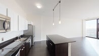 This modern single-floor unit showcases a clean, contemporary design characterized by crisp white walls and rich dark hardwood flooring that flows consistently throughout the space. The open-concept kitchen serves as a central highlight, featuring a large central island with a dark stone countertop, white upper cabinetry that extends to the ceiling for generous storage, dark lower cabinets, and a full suite of stainless steel appliances including a refrigerator, electric range, microwave, and dishwasher. Pendant lighting above the island and a track lighting system add both functionality and style to the space. The living and bedroom areas are bathed in natural light courtesy of expansive floor-to-ceiling windows, which frame sweeping city views and create a bright, airy ambiance throughout. The bedroom offers direct access to a generously sized walk-in closet equipped with dual wire shelving rods along multiple walls, providing ample storage. A dedicated laundry room houses a stacked washer and dryer unit, adding convenience to daily living. The bathroom is finished with large-format grey tile flooring, dark cabinetry, and a white countertop vanity, maintaining the cohesive modern aesthetic found throughout the rest of the unit.