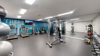 Located at 120 N Frontage Rd in Mansfield Center, CT, this fitness center offers a comprehensive workout facility across a single floor. The space is thoughtfully designed to accommodate a wide range of exercise needs, with durable rubber flooring throughout the main workout area providing both safety and comfort.

The primary exercise area is highlighted by a bold teal accent wall featuring motivational text, a mounted flat-screen TV, and a full-length mirror that visually expands the space. This zone houses a multi-functional cable machine, a selection of stationary bikes, treadmills, and additional cardio equipment. Overhead, recessed linear LED lighting ensures the area is brightly and evenly illuminated.

A dedicated free weights section includes a well-stocked dumbbell rack and a tiered stability ball stand, set against clean white walls with ample open floor space for functional training. An adjacent hallway area transitions into a secondary room with light wood-tone flooring, suitable for stretching, yoga, or low-impact activities.

The facility maintains a clean, modern aesthetic with white walls, low ceilings with flush lighting, and a practical layout that maximizes usable floor space. The overall ambiance is functional and energizing, making it a fully equipped destination for cardiovascular, strength, and flexibility training.