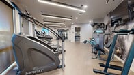 Located at 222 W Boston St in Chandler, Arizona, this modern fitness center offers a well-appointed, single-floor layout designed to support a comprehensive workout experience. The space features light wood-look flooring throughout, paired with crisp white walls and a clean, contemporary aesthetic that creates an open, energizing atmosphere.

Overhead, a combination of linear LED strip lighting and recessed fixtures provides bright, even illumination across the entire floor. Natural light is introduced through multiple windows positioned along the perimeter walls, offering views of the surrounding Chandler skyline and enhancing the overall sense of openness.

The cardio zone is well-equipped with a row of Spirit-brand stair climbers, treadmills, and stationary bikes, each outfitted with digital display consoles. The strength training area includes a multi-functional cable machine, a Smith machine with an adjustable bench, and a tiered dumbbell rack stocked with a full range of free weights. A wall-mounted flat-screen television and a bold, industrial-style accent wall mural add visual interest to the strength zone.

A restroom and vanity area is accessible directly from the main floor, adding convenience for users of the facility. The overall design balances functionality with a polished, modern finish, making this fitness center a practical and well-equipped amenity.