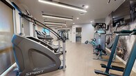 Located at 222 W Boston St in Chandler, Arizona, this modern fitness center offers a well-appointed, single-floor layout designed to support a comprehensive workout experience. The space features light wood-look flooring throughout, paired with crisp white walls and a clean, contemporary aesthetic that creates an open, energizing atmosphere.

Overhead, a combination of linear LED strip lighting and recessed fixtures provides bright, even illumination across the entire floor. Natural light is introduced through multiple windows positioned along the perimeter walls, offering views of the surrounding Chandler skyline and enhancing the overall sense of openness.

The cardio zone is well-equipped with a row of Spirit-brand stair climbers, treadmills, and stationary bikes, each outfitted with digital display consoles. The strength training area includes a multi-functional cable machine, a Smith machine with an adjustable bench, and a tiered dumbbell rack stocked with a full range of free weights. A wall-mounted flat-screen television and a bold, industrial-style accent wall mural add visual interest to the strength zone.

A restroom and vanity area is accessible directly from the main floor, adding convenience for users of the facility. The overall design balances functionality with a polished, modern finish, making this fitness center a practical and well-equipped amenity.