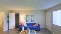 This single-story space presents a straightforward and functional layout centered around a living area that serves as the focal point of the home. The living room is finished with light gray tile flooring, which provides a clean and low-maintenance foundation throughout the space. Neutral, muted wall tones create a calm backdrop that allows furnishings and décor to stand out, as illustrated by the bold blue tufted sofa positioned against the main wall. A textured popcorn ceiling adds a subtle dimensional quality to the overhead finish. The room benefits from ambient lighting, with a floor lamp providing a warm glow to the space. A closet is visible to one side, offering accessible built-in storage. The overall layout appears open and uncluttered, lending the area a sense of spaciousness. The minimalist wall treatment, accented by a pair of abstract canvas artworks in cool blue and gold tones, contributes to the space's understated aesthetic. The design strikes a balance between simplicity and livability, making it a versatile environment suitable for a range of personal styles and furnishing arrangements.