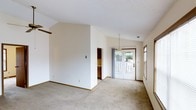 This single-floor space is characterized by its open, airy layout and vaulted ceilings that contribute to a sense of spaciousness throughout the main living area. The interior is finished in soft, neutral tones with cream-colored walls and light beige carpeting underfoot, creating a clean and versatile backdrop. A textured ceiling adds subtle visual interest overhead.

Natural light filters into the space through a set of glass French doors at the rear, which open directly to an outdoor deck or patio area, offering views of surrounding greenery. A sidelight window near the entrance further enhances brightness within the space. A pendant light fixture is positioned near the dining or transitional area, providing focused overhead illumination.

A partial wall divides the main living area from an adjoining room, which appears to feature cabinetry with wood-toned finishes, suggesting a kitchen or utility space beyond. Warm wood trim accents the doorframes and baseboards throughout, complementing the neutral palette. The overall ambiance is calm and understated, offering a functional and adaptable living environment ready for personalization.