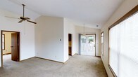 This single-floor space is characterized by its open, airy layout and vaulted ceilings that contribute to a sense of spaciousness throughout the main living area. The interior is finished in soft, neutral tones with cream-colored walls and light beige carpeting underfoot, creating a clean and versatile backdrop. A textured ceiling adds subtle visual interest overhead.

Natural light filters into the space through a set of glass French doors at the rear, which open directly to an outdoor deck or patio area, offering views of surrounding greenery. A sidelight window near the entrance further enhances brightness within the space. A pendant light fixture is positioned near the dining or transitional area, providing focused overhead illumination.

A partial wall divides the main living area from an adjoining room, which appears to feature cabinetry with wood-toned finishes, suggesting a kitchen or utility space beyond. Warm wood trim accents the doorframes and baseboards throughout, complementing the neutral palette. The overall ambiance is calm and understated, offering a functional and adaptable living environment ready for personalization.