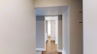 This single-story space presents a simple, well-maintained interior defined by a neutral color palette of soft grays and warm whites. The hallway visible in the image offers a glimpse into the layout, showcasing wide doorway openings with clean white trim that frame the passage between rooms. The wood-look flooring runs continuously through the corridor, adding warmth and visual cohesion to the space. Walls are finished in a muted gray tone, complemented by white baseboards that provide a crisp contrast. Natural light filters in through a window visible at the end of the hallway, brightening the interior and contributing to an open, airy feel. The straightforward layout and understated design choices create a functional and livable environment. The wide doorway openings enhance the sense of flow between spaces, making movement throughout the unit feel effortless. Overall, the space reflects a clean, modern aesthetic with practical finishes suited for everyday living.