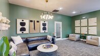 Located at 300 Gatehouse Lane in Odenton, Maryland, this single-floor lounge space offers a thoughtfully curated interior that blends modern aesthetics with a welcoming atmosphere. The space is defined by its rich sage green walls, which set a calm and sophisticated tone throughout.

The main lounge area serves as the centerpiece, anchored by a generous dark leather sectional sofa paired with an elegant round white marble coffee table featuring a gold-trimmed tray. Above, a striking multi-bulb chandelier with a brushed metal ring frame provides warm, ambient lighting. A triptych of bold abstract artwork adorns the feature wall, complemented by sculptural paper art installations on an adjacent wall, adding layers of visual interest.

Patterned carpet tile in muted grey tones runs consistently throughout, grounding the space with texture and continuity. A secondary seating area features two upholstered armchairs in a neutral stripe fabric, arranged alongside a petite accent table and a gallery wall of framed neutral art panels. Natural light filters in through white-trimmed windows, brightening the space further.

Additional utility is provided by a console table near the entry area, accented with decorative objects and a potted plant. Frosted glass interior doors offer privacy while maintaining an open, airy feel. Overall, the space conveys a polished, contemporary character ideal for communal gathering or relaxation.