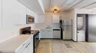 This single-floor studio-style unit showcases an industrial-modern aesthetic defined by polished concrete floors throughout and a clean, open layout. The main living area is generously proportioned, featuring exposed ceiling ductwork, a ceiling fan with an integrated mini-split system for climate control, and a row of windows offering views of the surrounding urban skyline. Natural light filters in across the reflective concrete floors, enhancing the sense of openness.

The kitchen is well-appointed with a full suite of stainless steel appliances, including a refrigerator, electric range with oven, over-the-range microwave, and dishwasher. Flat-front cabinetry in a light grey-white finish extends along the walls, paired with white countertops for a cohesive, contemporary look. Track lighting accents the upper cabinetry area.

The full bathroom is functionally designed with a white vanity featuring an undermount sink, a large mirror, and a tub-shower combination enclosed by white surround panels. Chrome fixtures and a nautical-style flush-mount ceiling light complete the space. The consistent use of polished concrete flooring ties the bathroom to the rest of the unit, reinforcing the space's modern, loft-inspired character.