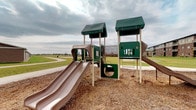 This outdoor playground offers a thoughtfully designed recreational space ideal for active play. The centerpiece is a multi-tower play structure finished in green and tan tones, featuring distinctive green shingled rooftops that provide shade and visual character. The structure includes dual slides, tunnel crawl-throughs, climbing panels, and rope net elements, offering a variety of engaging activities for different skill levels. The play area is surfaced with a generous layer of wood chip mulch, providing a cushioned, safety-conscious ground cover beneath and around all equipment. Surrounding the structure, the landscape opens up into a well-maintained grassy lawn, giving ample room to run and explore beyond the play equipment. Curved paved pathways wind through the park, connecting the playground to the broader grounds and encouraging easy navigation throughout the space. The open, expansive setting provides wide sightlines across the area, with the surrounding neighborhood visible in the distance. The overall design balances functionality and safety, making this a welcoming community recreation space suited for children of varying ages.
