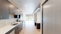 Located in Iowa City, IA, this compact yet thoughtfully designed single-floor unit offers a streamlined open layout that maximizes every square foot. Light-toned hardwood flooring flows throughout, lending warmth and visual continuity across all areas of the space.

The kitchen is a standout feature, outfitted with rich dark-walnut cabinetry extending to the ceiling for ample storage, white quartz countertops, a classic subway tile backsplash, and a full suite of stainless steel appliances including a refrigerator, electric range, microwave, and dishwasher. The galley-style configuration keeps the workspace efficient and well-organized.

The living area is bright and airy, anchored by floor-to-ceiling sliding glass doors that open to an outdoor balcony, inviting abundant natural light and outdoor views. An exposed cylindrical ductwork detail adds an industrial-modern architectural accent to the space. A built-in desk or counter nook provides a dedicated workspace or dining area.

The bathroom is finished with large-format grey marble-style wall tiles in a glass-enclosed shower/tub combination, creating a clean and polished aesthetic. Throughout the unit, dark wood trim provides a striking contrast against the crisp white walls, contributing to a cohesive contemporary design.