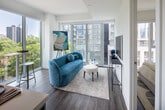 Located in Toronto's dynamic Church-Wellesley Village, Suite 601 at 8 Gloucester Street is a thoughtfully designed two-bedroom, two-bathroom residence that makes excellent use of its compact footprint. The open-concept living area is defined by expansive floor-to-ceiling windows on two sides, flooding the space with natural light and offering striking views of the surrounding cityscape and tree canopy below. Hardwood-style flooring runs throughout, tying the rooms together cohesively.

The kitchen is a functional centerpiece, featuring warm wood-grain cabinetry extending to the ceiling, white quartz countertops, stainless steel appliances, and a peninsula with bar seating — ideal for casual dining. Track lighting above adds a polished, modern touch.

Both bedrooms are bright and well-proportioned. The primary bedroom benefits from a large sliding glass door that opens to city views, while the second bedroom maintains the same clean, minimal aesthetic. Each room is served by its own bathroom: one featuring a glass-enclosed walk-in shower with floor-to-ceiling grey tile, and the other a full soaker tub with matching wood-toned vanity cabinetry. Throughout the unit, the design palette of white walls, warm wood accents, and light tile creates a clean, contemporary ambiance.