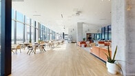 This single-floor space is a thoughtfully designed communal amenity level that blends contemporary architecture with a welcoming, hospitality-inspired atmosphere. The most striking architectural feature is the continuous wall of floor-to-ceiling glass that runs along the perimeter, flooding the interior with abundant natural light and framing panoramic city skyline views. Light wood-toned tile flooring extends throughout, creating a warm, cohesive foundation that ties the various zones together seamlessly.

The space is divided into several distinct yet open areas. A café station anchors one section, featuring a terrazzo-accented counter, pendant lighting, digital menu displays, and a fully equipped service bar. Adjacent to this is a casual dining zone furnished with lightweight wooden chairs and café-style tables positioned to take advantage of the natural light streaming through the expansive windows.

A lounge area provides plush, terracotta-toned sectional sofas and an assortment of accent chairs arranged on patterned carpeting, offering a comfortable setting for relaxed conversation or casual work. A separate seating section, elevated by oversized dome pendant lights, presents a brighter, airier feel with views directly overlooking the cityscape. Sculptural art installations and exposed concrete columns add visual interest and an industrial-modern character throughout the space.