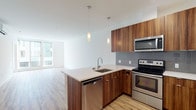 This contemporary single-floor unit offers a thoughtfully designed open-concept layout that seamlessly connects the kitchen and living areas. The kitchen is a standout feature, showcasing rich wood-grain cabinetry that extends to the ceiling, a grey tile backsplash, and a full suite of stainless steel appliances including a refrigerator, electric range with oven, built-in microwave, and dishwasher. A peninsula with a white quartz countertop and an undermount sink serves as both a functional workspace and a natural divider between the kitchen and living space. Elegant pendant lights hang above the peninsula, adding warmth to the clean, modern aesthetic. Light hardwood flooring flows consistently throughout the entire unit, tying the spaces together cohesively. The living area is bright and open, benefiting from expansive floor-to-ceiling windows that invite an abundance of natural light and provide views of the surrounding exterior. A wall-mounted mini-split HVAC unit ensures year-round climate comfort. The bedroom is generously proportioned, featuring large windows, a closet, and a neutral grey accent wall. The bathroom is finished with a double undermount sink set into a marble-look quartz vanity, complemented by a large mirror and clean, minimal fixtures. The overall ambiance is modern, clean, and move-in ready.