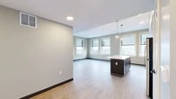 Located in the heart of downtown Utica, NY, this thoughtfully designed two-bedroom, two-bathroom residence offers a contemporary open-concept layout across a single floor. The combined kitchen and living area serves as the centerpiece of the home, featuring light hardwood-style flooring, recessed lighting, and large windows that flood the space with natural light while framing elevated views of the Utica skyline. The kitchen is well-appointed with dark cabinetry, a white subway tile backsplash, a large central island with a light quartz countertop, elegant pendant lighting, and a full suite of stainless steel appliances. The two bedrooms each benefit from multiple windows offering sweeping city views — one features plush carpeting and soft sage-toned walls, while the primary bedroom provides a generous footprint with hard-surface flooring and warm neutral tones. Three closets throughout the unit offer ample storage. The bathrooms are finished with large-format tile flooring and stylish vanities, including a standout blue vanity with a dark stone countertop. A hallway connects the private areas of the home seamlessly. The overall ambiance is clean, modern, and bright, with a strong sense of urban connectivity.