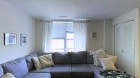 This single-floor living space centers around a generously sized gray sectional sofa, which serves as the focal point of the room and provides ample seating. The sofa is dressed with an assortment of decorative pillows in neutral tones and soft botanical prints, adding texture and a cohesive visual interest to the space. A large window positioned behind the sofa allows natural light to filter into the room, fitted with horizontal blinds for light control and framed by sheer white patterned curtains that extend across the wall, lending an airy, soft aesthetic. The walls are finished in a clean, neutral tone that complements the understated color scheme throughout. A small framed piece of wall art is visible to the right, contributing a subtle decorative touch without overwhelming the space. The ceiling features a central HVAC vent, indicating a built-in climate control system. The overall ambiance is calm and functional, with a casual, lived-in comfort that makes the space welcoming. The design choices emphasize simplicity and practicality, making it a versatile area well-suited for relaxing or entertaining.