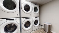 This laundry room is designed for shared use and is well-equipped to handle high-volume laundry needs. The room features multiple Whirlpool front-load washers and dryers arranged in stacked configurations along one wall, maximizing the use of vertical space while keeping the floor area open and accessible. Each unit offers modern digital controls, providing a range of wash and dry cycle options for users.

The flooring throughout the room consists of wood-look vinyl planks in a warm gray-brown tone, lending a clean and contemporary appearance. Neutral gray walls and white baseboards contribute to a tidy, utilitarian aesthetic. A built-in folding table with a light wood surface is positioned along one wall, providing a practical workspace for sorting and folding laundry. A stainless steel step-on trash can is conveniently placed nearby.

The room is accessed through a door featuring a glazed upper panel and a brushed stainless steel kick plate, with a keyed lever handle suggesting secured access. A textured drop ceiling completes the space. Overall, the laundry room is functional, efficiently organized, and maintained with a clean, modern finish.
