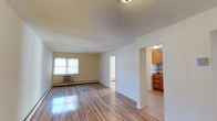 Maple Crest, located at 780 Rockaway Avenue in Valley Stream, NY, presents a well-maintained single-floor residence designed for comfortable everyday living. The open living area is immediately welcoming, featuring gleaming hardwood floors that reflect natural light streaming through a large window fitted with bright vertical blinds. The neutral, warm-toned walls create a clean and airy atmosphere throughout. A baseboard heating system and a wall-mounted air conditioning unit ensure year-round climate comfort. The living space flows seamlessly into the adjacent kitchen, accessible through an open arched passageway. The kitchen is outfitted with warm wood-toned cabinetry, a tile backsplash, and light tile flooring, offering a practical and tidy cooking environment. A doorway visible from the main living area leads to additional rooms, suggesting a thoughtfully laid-out floor plan that balances openness with privacy. The overall ambiance is bright, clean, and move-in ready, with a straightforward design that emphasizes functionality and ease of living. Maple Crest's Valley Stream location provides convenient access to local amenities, transportation, and the broader Long Island community.