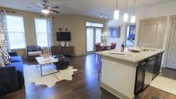 This well-appointed single-floor residence showcases an open-concept layout that seamlessly connects the kitchen, dining, and living areas into one cohesive and inviting space. The kitchen anchors the layout with a granite-topped island that doubles as a breakfast bar, offering both functionality and style. Pendant lighting hangs above the island, adding warm, focused illumination that complements the track lighting overhead.

Rich, dark hardwood flooring flows continuously throughout the space, grounding the interior and lending a sense of warmth and sophistication. The living area is bright and airy, benefiting from multiple windows dressed with white horizontal blinds and patterned curtains that allow ample natural light to filter in. A ceiling fan overhead adds comfort, while the open arrangement provides generous room for furnishings.

The dining area sits naturally between the kitchen and living room, framed by additional windows that further enhance the sense of openness. A bedroom is visible in the background, hinting at a thoughtfully connected floor plan. Neutral wall tones provide a versatile backdrop throughout, making the space feel both welcoming and easy to personalize. Overall, the layout balances practicality with a contemporary aesthetic.
