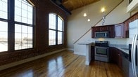 This two-story loft blends industrial character with modern comfort, offering one bedroom, one bathroom, and a lofted den. The main living area makes an immediate impression with soaring ceilings clad in warm wood paneling and dramatic exposed brick walls that speak to the building's historic roots. A large arched warehouse-style window floods the space with abundant natural light while framing sweeping views of the surrounding cityscape. Rich hardwood floors run throughout, adding warmth and continuity to the open layout. Exposed ductwork and structural beams overhead reinforce the authentic industrial aesthetic. The kitchen is positioned adjacent to the living area and features stainless steel appliances, including a range and built-in microwave, dark wood cabinetry, and a granite-topped peninsula that provides additional counter space and casual seating. Pendant lighting and a track light system accent the kitchen and living zones. A staircase leads to the upper level, where the lofted den offers a versatile secondary space ideal for a home office, reading nook, or lounge area. The two-level layout provides a sense of openness and dimension rarely found in urban residences of this size, making it a distinctive and thoughtfully designed living space.