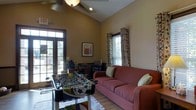 This single-story recreational lounge offers a welcoming and casual atmosphere designed for leisure and social interaction. The room features vaulted ceilings that enhance the sense of openness, complemented by a ceiling fan that adds to the comfortable environment. Large French doors along one wall flood the space with natural light, providing a view of the outdoors, while an additional window dressed with patterned curtains and blinds adds further brightness to the warm, tan-colored walls.

The centerpiece of the room is a foosball table positioned on a colorful area rug over light hardwood-style flooring, making it an inviting focal point for recreation. A plush red sofa with decorative throw pillows offers comfortable seating nearby, ideal for relaxing between games. Along the back wall, a small entertainment or refreshment station is visible, suggesting the space is equipped to support extended social gatherings. A framed piece of wall art adds a decorative touch to the overall design. The combination of recreational amenities, natural light, and relaxed furnishings creates a casual, community-oriented atmosphere well-suited for group activities and socializing.
