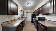 This two-story home offers three bedrooms and two bathrooms distributed across two floors of comparable size. The main floor houses the kitchen and living room, while the upper floor accommodates the bedrooms and remaining bathrooms, connected by a central hallway.

The galley-style kitchen is a standout feature, finished with rich dark espresso cabinetry extending to the ceiling and topped with granite-look countertops. A double-basin sink sits beneath a window, allowing natural light into the space. Black appliances, including an electric range, refrigerator, and dishwasher, complement the cabinetry. A textured ceiling and wood-look flooring run throughout the kitchen and extend into the living area.

The living room features the same wood-look flooring, a ceiling fan with light fixture, and a built-in niche, providing a functional accent wall detail. Neutral warm-beige walls create a cohesive, understated palette throughout.

The bedrooms feature plush carpeting and ceiling fans, offering comfort and practicality. The primary bedroom includes closet space with dedicated storage. Hallway access connects the upper-level rooms efficiently, and the two closets provide additional organizational utility throughout the home.