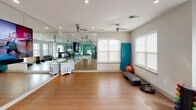 Located at 12025 Landing Place Lane in Charlotte, NC, this single-floor fitness facility offers a thoughtfully designed environment suited for a wide range of workout activities. The main exercise area features smooth hardwood-style flooring that provides an ideal surface for group fitness, yoga, stretching, and cardio training. A full wall of floor-to-ceiling mirrors visually expands the space while allowing users to monitor their form during workouts. A large flat-screen display is mounted prominently on the mirrored wall, enabling virtual workout sessions and instructional programming.

Ceiling fans are installed throughout, promoting air circulation during active sessions, while recessed lighting ensures the space remains bright and well-illuminated. Multiple windows along the exterior walls allow generous natural light to fill the room and provide views of the surrounding neighborhood.

An adjacent glass-walled weight room offers a distinct area for strength training, housing a variety of equipment including dumbbells, medicine balls, and cardio machines. This transparent enclosure maintains visual connectivity with the main studio. Additional fitness accessories, including kettlebells, resistance tools, exercise mats, and step platforms, are organized throughout the space, making it a comprehensive and functional fitness destination.