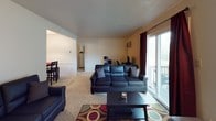 This single-story, one-bedroom unit offers a practical and functional layout across several well-defined areas. The galley-style kitchen features dark wood cabinetry lining both walls, providing ample storage space. White appliances, including a refrigerator, electric range, and dishwasher, are complemented by a double stainless steel sink and patterned backsplash. Tile-style flooring and a door at the far end of the kitchen add to its efficient design.

The living area is carpeted and open, connecting seamlessly to a dining space. Large sliding glass doors allow natural light to fill the room and provide direct access to an outdoor balcony with a wooden railing. The living room also features multiple doorways leading to other areas of the unit, including a second sitting or flex space.

The bedroom is carpeted and includes a textured ceiling, a window fitted with a wall air conditioning unit, and two interior doors offering closet or additional storage access. The full bathroom includes a vanity with a mirrored medicine cabinet, lighted mirror, toilet, and a shower-tub combination enclosed by a decorative fabric curtain.