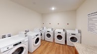 Located at 2961 Lenora Church Rd in Snellville, Georgia, this dedicated laundry center is a well-appointed, single-floor facility designed to serve its users efficiently. The room is finished with warm hardwood-style flooring and neutral, light-toned walls that create a clean and welcoming atmosphere.

The space is equipped with multiple front-loading washing machines and dryers arranged along the perimeter walls, providing ample capacity for simultaneous use. The machines are uniformly white and neatly aligned, contributing to the room's organized appearance. Each unit is positioned on a raised platform with individual utility hookups, ensuring convenient and reliable operation.

A standout feature of the space is a large folding table with a granite-style countertop, supported by a warm wood base. This generous surface provides ample room for sorting and folding laundry. A window fitted with a blind allows natural light to filter into the room while maintaining privacy.

The facility also displays posted laundry center rules, reinforcing a community-oriented and orderly environment. Recessed lighting and standard overhead fixtures provide consistent illumination throughout. The overall layout is thoughtfully designed to maximize functionality, making this laundry center a practical and well-maintained shared resource.