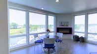 This single-floor space offers a thoughtfully designed interior with a bright, contemporary aesthetic. The dining area is a standout feature, bathed in natural light streaming through expansive floor-to-ceiling windows that wrap around the corner of the room, framing stunning panoramic views of a marina filled with sailboats and lush greenery. The light hardwood flooring complements the clean, neutral wall tones, creating an airy and welcoming atmosphere throughout.

A sleek, wall-mounted electric fireplace with ambient accent lighting serves as a striking focal point in the living and dining area, adding both warmth and a modern design element. Above it, artwork echoes the nautical theme of the surrounding waterfront environment.

The dining setup features a round table with wicker-style chairs accented by vibrant blue cushions, adding a pop of color that ties in beautifully with the marina backdrop. Recessed ceiling lighting provides even illumination throughout the space, complementing the natural light during evening hours.

Overall, this space strikes a balance between modern design and natural surroundings, with its marina-facing windows, contemporary fireplace, and clean finishes creating an inviting environment that highlights its exceptional waterfront setting.