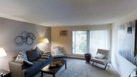 This single-floor residence features a welcoming living room that serves as the heart of the space. The room is finished with wall-to-wall carpeting in a neutral tone, contributing to a warm and comfortable atmosphere. A large sliding glass door dominates one wall, flooding the interior with natural light and providing direct access to a private balcony that overlooks a green, tree-lined outdoor area. Vertical blinds offer privacy and light control as needed. The living room comfortably accommodates a dark upholstered sofa paired with accent chairs, centered around a dark wood coffee table. A wall-mounted air conditioning unit ensures year-round climate control, while a baseboard heating system runs along the exterior wall. Soft ambient lighting from a floor lamp complements the natural light. Subtle wall décor adds a personal touch without overwhelming the clean, understated aesthetic. The overall ambiance is practical and inviting, with a layout that maximizes the sense of openness while maintaining a cozy, livable feel suited for everyday comfort.