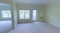 This single-floor space offers a clean, understated aesthetic defined by soft, neutral wall tones and plush wall-to-wall carpeting that create a calm and welcoming atmosphere throughout. The main living area is generously sized, providing an open and airy feel well-suited for a variety of furniture arrangements and living styles. A large double-pane window anchors one wall, flooding the interior with abundant natural light while framing views of a greenery-lined outdoor area. Adjacent to the window, a glass-panel door offers direct access to the exterior, enhancing both the flow of natural light and the sense of connection to the outdoors. Crown molding lines the ceiling, adding a subtle architectural refinement to the otherwise minimalist design. Practical elements are thoughtfully integrated, including wall-mounted climate control units and ventilation grilles that ensure year-round comfort. The white baseboards and trim provide a crisp contrast against the light walls, contributing to the space's tidy and well-maintained appearance. Overall, this unit presents a bright, functional, and move-in-ready environment with a straightforward layout that balances comfort with practicality.
