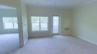 This single-floor space offers a clean, understated aesthetic defined by soft, neutral wall tones and plush wall-to-wall carpeting that create a calm and welcoming atmosphere throughout. The main living area is generously sized, providing an open and airy feel well-suited for a variety of furniture arrangements and living styles. A large double-pane window anchors one wall, flooding the interior with abundant natural light while framing views of a greenery-lined outdoor area. Adjacent to the window, a glass-panel door offers direct access to the exterior, enhancing both the flow of natural light and the sense of connection to the outdoors. Crown molding lines the ceiling, adding a subtle architectural refinement to the otherwise minimalist design. Practical elements are thoughtfully integrated, including wall-mounted climate control units and ventilation grilles that ensure year-round comfort. The white baseboards and trim provide a crisp contrast against the light walls, contributing to the space's tidy and well-maintained appearance. Overall, this unit presents a bright, functional, and move-in-ready environment with a straightforward layout that balances comfort with practicality.