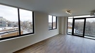 This single-floor space offers a clean, modern aesthetic defined by its open layout and abundant natural light. The living area is anchored by two oversized black-framed windows that frame expansive views of the surrounding cityscape, including streets, neighboring buildings, and the urban skyline beyond. The elevated position of the unit enhances these views, creating a strong sense of connection to the city below.

The flooring throughout is a warm-toned hardwood-style material that adds texture and visual warmth to the otherwise neutral interior. Walls are finished in a crisp, light beige tone that complements the natural light flowing in from multiple directions. The ceiling features a modern multi-directional track light fixture, providing both functional and accent lighting.

A set of dark-framed glass doors leads to an outdoor balcony, extending the living space and offering additional access to open-air city views. The minimalist design throughout keeps the focus on the architectural details and the panoramic urban backdrop, resulting in a bright, airy, and contemporary living environment.