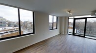 This single-floor space offers a clean, modern aesthetic defined by its open layout and abundant natural light. The living area is anchored by two oversized black-framed windows that frame expansive views of the surrounding cityscape, including streets, neighboring buildings, and the urban skyline beyond. The elevated position of the unit enhances these views, creating a strong sense of connection to the city below.

The flooring throughout is a warm-toned hardwood-style material that adds texture and visual warmth to the otherwise neutral interior. Walls are finished in a crisp, light beige tone that complements the natural light flowing in from multiple directions. The ceiling features a modern multi-directional track light fixture, providing both functional and accent lighting.

A set of dark-framed glass doors leads to an outdoor balcony, extending the living space and offering additional access to open-air city views. The minimalist design throughout keeps the focus on the architectural details and the panoramic urban backdrop, resulting in a bright, airy, and contemporary living environment.