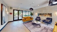 This contemporary lobby is designed to make a strong first impression, combining modern aesthetics with functional comfort. The space is anchored by a striking floor-to-ceiling stone accent wall housing a sleek linear fireplace, which serves as the focal point of the main seating area. Flanking wall sconces cast a warm, ambient glow against the textured stone, complementing the bold black fringe pendant light fixtures overhead.

The seating arrangement features rich amber leather sofas paired with deep navy accent chairs, centered around a marble-topped coffee table set atop a purple-toned area rug. Large-format light stone tile flooring runs throughout, creating a clean and cohesive aesthetic. Expansive black-framed glass doors and floor-to-ceiling windows flood the space with natural light and provide direct views of a landscaped outdoor courtyard.

Decorative circular wall panels and abstract artwork add artistic character along the corridors, which transition from the tiled lobby to carpeted hallways leading to individual units. Lush potted greenery punctuates the space, enhancing the welcoming atmosphere. Overall, the lobby balances contemporary design with inviting warmth, creating a polished and comfortable communal environment.