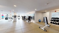 Located at 215 Grove St in Jersey City, NJ, this well-appointed fitness center offers a generous, open exercise space designed to accommodate a full range of workout routines. The room features a clean, modern aesthetic with white walls, recessed lighting, and large floor-to-ceiling windows that flood the space with natural light and provide views of the surrounding area.

The flooring throughout consists of durable, large-format rubber-backed tiles, providing both safety and comfort underfoot. Full-length wall mirrors are strategically positioned throughout the space, visually expanding the room and allowing users to monitor their form during workouts.

The cardio zone is equipped with multiple treadmills, elliptical trainers, and stationary bikes, including cycling-style bikes positioned near a dedicated mirrored alcove. The strength training area features a robust selection of selectorized machines, adjustable benches, and a well-stocked dumbbell rack with a full range of weights. Additional functional training accessories, including medicine balls, kettlebells, and resistance tools, are neatly organized along the walls.

The layout is thoughtfully zoned to allow simultaneous use of cardio and strength equipment without crowding. The combination of abundant natural light, mirrored walls, and a spacious floor plan creates a bright, motivating environment well-suited for a comprehensive fitness facility.