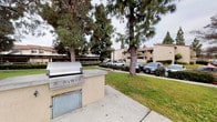 This single-floor community offers a thoughtfully designed outdoor environment with a variety of shared amenities. The grounds are anchored by mature pine trees that provide natural shade and greenery throughout the property. Concrete pathways wind through the landscaped areas, connecting key points of interest across the community.

A standout feature is the built-in outdoor BBQ station, constructed with a stucco finish and housing a stainless steel grill, making it well-suited for outdoor gatherings. Nearby, an open grassy lawn provides open recreational space, complemented by a dedicated bike rack for residents' convenience.

The swimming pool area serves as a central amenity, enclosed by a metal security fence and surrounded by patio furniture, including chairs and a table, offering a comfortable space for relaxation. Lush shrubs and flowering plants line the pool perimeter, enhancing the overall aesthetic.

The residential buildings feature a neutral stucco exterior with wood accents and balconies, blending cohesively with the landscaped surroundings. Mulched garden beds and trimmed hedges border the building foundations and walkways, contributing to the well-kept character of the community. Parking areas are conveniently accessible from the grounds.