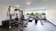 Located at 560 Proudfoot Ln in London, Ontario, this fitness facility offers a well-equipped exercise room designed to support a comprehensive workout experience. The space features dark rubber flooring throughout, providing durability and safety underfoot, while white walls and recessed ceiling lighting create a clean, functional atmosphere.

The room is outfitted with a variety of cardio machines, including treadmills, stationary spin bikes, and a rowing machine, all positioned to take advantage of the natural light streaming in through a large window overlooking greenery outside. For strength training, the facility includes a multi-function cable machine, an adjustable weight bench, and a well-stocked dumbbell and kettlebell rack with a full range of weights. Medicine balls on a dedicated stand add further versatility to workout options.

A wall-mounted flat-screen television provides entertainment during workouts. Multiple gray utility doors lead to adjacent utility and storage areas. A first aid station is also present, ensuring safety within the space. The overall layout is efficient and thoughtfully organized, making the most of the available footprint to accommodate a wide range of fitness activities in a practical, no-frills environment.
