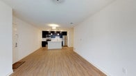 This well-designed single-floor space offers a bright and airy open-concept layout that seamlessly connects the living area to the kitchen. The light-toned hardwood-style flooring runs throughout, lending warmth and visual continuity to the entire space. Crisp white walls reflect light effectively, enhancing the clean and modern aesthetic.

The kitchen is a standout feature, showcasing dark espresso cabinetry that provides a striking contrast against the white walls and light countertops. A central island offers additional workspace and serves as a natural gathering point. Stainless steel appliances — including a refrigerator, electric range, and over-the-range microwave — add a polished, contemporary touch. A white subway tile backsplash completes the kitchen's refined look, while pendant lighting above the island provides focused, stylish illumination.

The open living area is generously proportioned, offering flexible space for various furniture arrangements. Recessed lighting and a ceiling-mounted fixture provide ample overhead illumination throughout. The layout flows intuitively, making the space feel connected and functional. Overall, the unit combines modern finishes with practical design for comfortable, everyday living.