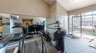 The Wood Creek Fitness/Spa is a single-story wellness facility designed to support both relaxation and active fitness. The space is divided into two distinct areas that together create a well-rounded amenity experience.

The spa area is a standout feature, housing an indoor whirlpool set beneath a dramatic vaulted ceiling with exposed wood beams and expansive floor-to-ceiling windows on multiple walls. The generous glazing floods the space with natural light and provides open views of the surrounding community and outdoor pool area. Stone tile surrounds the whirlpool, complementing the light blue walls and ceramic tile flooring, while wall sconces provide supplemental lighting. A glass-paneled door connects the spa to adjacent areas of the facility.

The fitness center is carpeted and equipped with a variety of cardio machines, including treadmills, ellipticals, and stationary bikes, as well as strength training equipment. Full-length mirrors line one wall, visually expanding the space, while a wall-mounted television provides entertainment during workouts. Vaulted ceilings contribute to an open, airy atmosphere, and windows along one side offer views of the outdoors, maintaining a bright and inviting environment throughout.