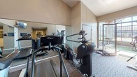 The Wood Creek Fitness/Spa is a single-story wellness facility designed to support both relaxation and active fitness. The space is divided into two distinct areas that together create a well-rounded amenity experience.

The spa area is a standout feature, housing an indoor whirlpool set beneath a dramatic vaulted ceiling with exposed wood beams and expansive floor-to-ceiling windows on multiple walls. The generous glazing floods the space with natural light and provides open views of the surrounding community and outdoor pool area. Stone tile surrounds the whirlpool, complementing the light blue walls and ceramic tile flooring, while wall sconces provide supplemental lighting. A glass-paneled door connects the spa to adjacent areas of the facility.

The fitness center is carpeted and equipped with a variety of cardio machines, including treadmills, ellipticals, and stationary bikes, as well as strength training equipment. Full-length mirrors line one wall, visually expanding the space, while a wall-mounted television provides entertainment during workouts. Vaulted ceilings contribute to an open, airy atmosphere, and windows along one side offer views of the outdoors, maintaining a bright and inviting environment throughout.