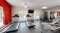 Located in Gulf Shores, Alabama, this fitness center offers a well-equipped exercise space designed to support a variety of workout routines. The room features a bold, energizing aesthetic defined by a vibrant red accent wall and a large typographic motivational mural that spans one full wall, creating a dynamic and inspiring atmosphere for users.

The space is outfitted with a comprehensive selection of equipment, including multiple treadmills, elliptical cross-trainers, a stationary bike, and a multi-station cable strength machine, providing options for both cardiovascular and resistance training. A wooden bench offers a convenient spot for stretching or rest between sets.

Durable rubber flooring covers the entire exercise area, providing traction and impact absorption suitable for high-intensity use. A full-length mirror along one wall visually expands the space and allows users to monitor their form during workouts.

Natural light filters in through several large windows, including a prominent window on the red accent wall and additional windows overlooking what appears to be a pool area, giving the room an open, airy feel. Overhead fluorescent lighting and a ceiling fan ensure the space remains bright and well-ventilated. A water dispenser is conveniently positioned along the wall, and a wall-mounted television provides entertainment during cardio sessions.