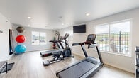 Located at 4451 Portrush Rd in Rapid City, South Dakota, this fitness center offers a clean, modern environment designed to support a comprehensive workout routine. The single-floor space features light wood-style flooring, crisp white walls, and recessed lighting throughout, creating a bright and welcoming atmosphere.

The cardio zone is equipped with Star Trac treadmills, an elliptical trainer, and a rowing machine, all positioned to take advantage of the natural light streaming in through multiple large windows. Views of the outdoor pool area and surrounding grounds are visible from the windows, adding an open, airy feel to the space.

A wall-mounted flat-screen television provides entertainment during workouts. The strength training area includes a well-stocked dumbbell rack with kettlebells, an adjustable weight bench, and a large floor-to-ceiling mirror that visually expands the room and allows for form checks during exercise.

Additional amenities include a stability ball rack stocked with multiple sized balls, wall-mounted exercise mats, a coat hook station, and industrial fans for ventilation. The layout is thoughtfully organized to allow ample floor space for stretching and floor exercises. The overall ambiance is functional yet comfortable, making this fitness center a well-rounded facility for residents seeking convenient on-site wellness amenities.