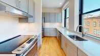 Situated in the heart of Providence's downtown on Westminster Street, this single-floor unit presents a clean, contemporary design characterized by warm hardwood flooring, white walls, and a bright, open aesthetic throughout. The layout, as seen in the floor plan, is well-organized with multiple rooms connected by a central corridor.

The main living area is generously proportioned, featuring track lighting overhead and large windows that frame sweeping views of Providence's urban skyline and historic architecture, flooding the space with natural light. Additional rooms, suitable for use as bedrooms or offices, continue the hardwood flooring and include ample window placement to ensure consistent natural light and city views.

The kitchen is well-appointed with gray shaker-style cabinetry, white countertops, stainless steel appliances including a range, dishwasher, and refrigerator, and a window overlooking the streetscape. Two bathrooms serve the unit: one features a tiled walk-in shower with a vanity and gray cabinetry, while the second includes a full soaking tub and shower combination with neutral tile flooring. Both bathrooms maintain a clean, modern finish consistent with the overall design of the space.